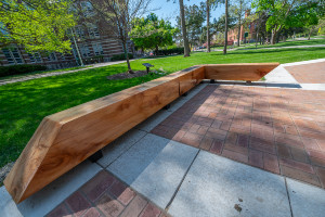 Bench made of solid wood in an L shape on either side of a brick walkway surrounded by green grass.